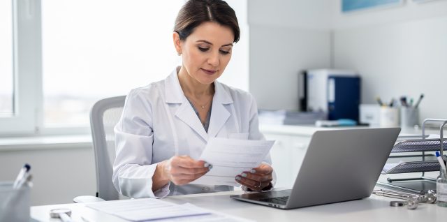 Médecin libérale dans un cabinet lumineux, consultant des documents comptables non lisibles à côté d’un ordinateur portable.